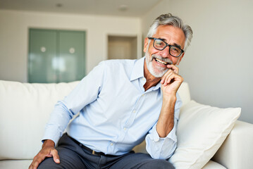 A 60 year old smiling positive business man with gray hair and beard and glasses sits comfortably on a sofa his chin resting on his hand.