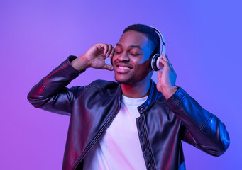 Music Lover. Handsome Black Guy In Wireless Headphones And Leather Jacket Listening His Favorite Songs, Relaxed African American Man Standing With Closed Eyes Under Neon Light Over Purple Background