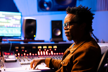 Fototapete - African american record producer working on mixing tunes in music studio, editing sounds in control room with technical equipment and console. Skilled audio expert producing new songs.