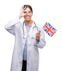 Wall Mural - Young hispanic doctor woman holding flag of united kingdom with happy face smiling doing ok sign with hand on eye looking through fingers