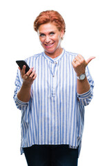 Wall Mural - Atrractive senior caucasian redhead woman texting using smartphone over isolated background pointing and showing with thumb up to the side with happy face smiling