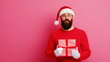 © Wonderful Studio - A man in a Santa hat and red sweater holds a gift box against a pink background, spreading holiday cheer.