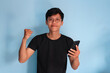 © Lailar Studio - Young Asian man smiling clenching his fist while holding a cell phone
