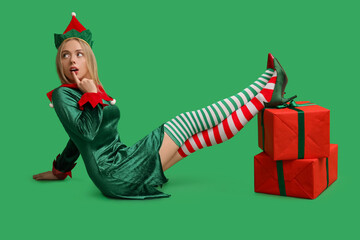 Sticker - Emotional young woman in elf costume with Christmas gift boxes on green background