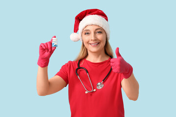 Wall Mural - Mature female doctor in Santa hat with jar of pills showing thumb-up gesture on blue background