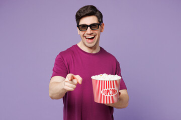 Poster - Young Caucasian man wear violet t-shirt casual clothes 3d glasses watch movie film hold bucket of popcorn in cinema point index finger camera on you isolated on pastel light purple background studio.