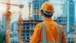 © ItziesDesign - Construction worker in orange uniform observing building site with cranes and skyscrapers in the background.