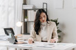 © Wicitr - Focused and Determined: A professional woman in a sleek beige blazer, intently reviewing financial documents in a modern home office setting.