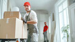 © Anoo - Professional movers transporting heavy boxes while a client in overalls and a red cap supervises in a light, calm room with white walls and furniture