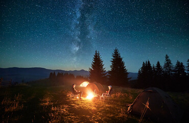 Wall Mural - Silhouette of couple hikers resting by campfire under star-filled sky, near illuminated tents at campsite. Night camping in mountains, night sky create magical and peaceful atmosphere.