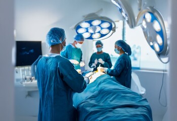 A surgeon in a blue surgical gown and cap is in an operating room. They are performing surgery while two other surgeons assist them.