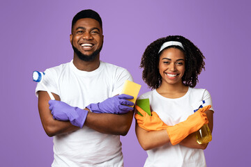 On Guard Of Cleanliness. Smiling black couple holding cleaning tools and posing with folded arms over pink studio background, free space