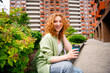 © deagreez - Joyful young redhead woman enjoying coffee outdoors in a sunny day in the city with natural green surroundings