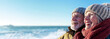 © David Giraud - Un couple de seniors heureux, souriant, sur la plage, portant des vêtements chauds lors d'une belle journée d'hiver, image avec espace pour texte.