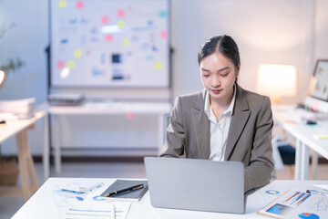 Young professional is concentrating on her laptop, surrounded by charts and documents, demonstrating the demanding nature of corporate life