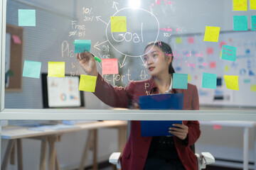 Businesswoman is analyzing business growth and planning a business strategy using adhesive notes on a glass wall in the office