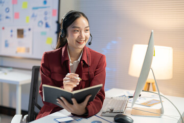 Asian businesswoman happily working late in her well-lit office, smiling during a video call, surrounded by technology and paperwork, showcasing her dedication to her career