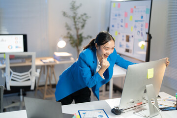 Young businesswoman working late at her office, celebrating success with a big smile, focused on her computer screen in a professional workspace filled with technology