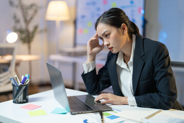 Young businesswoman is feeling tired and having a headache while working on a laptop late at night. She is sitting at her desk in a modern office