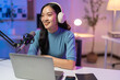 © Tj - Young asian woman is recording a podcast from home, talking into a microphone and using a laptop. She is smiling and looking away from the camera