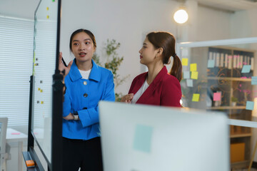 Two businesswomen having a discussion in the office while pointing at a glass whiteboard with sticky notes