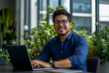 A smiling Hispanic man sitting at a desk with a laptop, looking into the camera, with a modern office background.