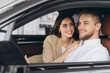 © anatoliycherkas - Portrait of smiling beautiful couple sitting in modern luxury car, happy excited young man and woman buying new car in dealership.