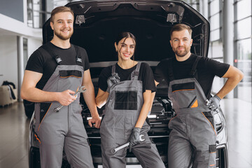 Three smiling workers, mechanics two men and woman in grey uniform standing at open hood, garage vehicle shop, auto mechanic technician maintenance customer car automobile at repair service.