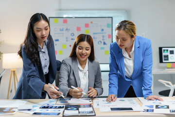 Wall Mural - Three asian businesswomen working together, leaning over a desk covered with financial charts and using markers to highlight key data