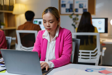 Young asian businesswoman is concentrating on her work on a laptop, surrounded by colleagues in a brightly lit office late at night