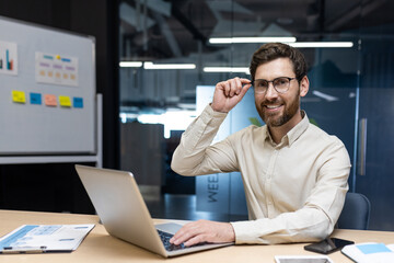 Portrait of a young smiling and successful young businessman sitting in the office at a desk with a laptop, holding glasses and confidently looking at the camera
