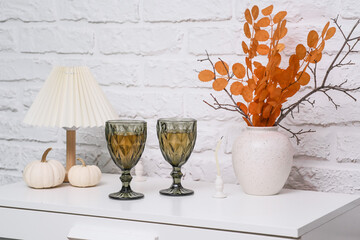 This stylish autumn decoration showcases two green glasses of wine, vase with autumn branches of yellow leaves, pumpkins and warm colors, creating a lovely fall ambiance.