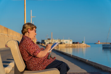 An older woman writing in a notebook, sitting on a bench near the sea. 