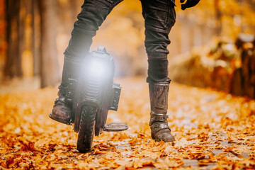 Detailed view of an electric unicycle wheel on an autumn forest path covered with orange leaves, highlighting the headlight and rider’s boots.