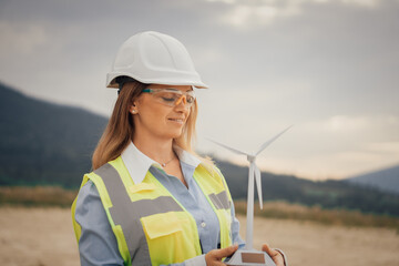 A young female engineer proudly showcases a model of a wind turbine at a renewable energy site, highlighting her dedication to sustainable development and innovation within the engineering field