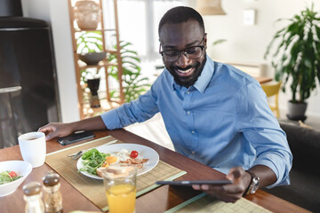 A smiling African-American man enjoys breakfast while using his tablet. He is seated at a table with a healthy meal and juice, in a cozy and modern home setting.