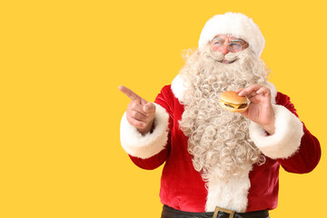 Canvas Print - Santa Claus with tasty burger pointing at something on yellow background