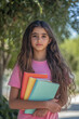 © AI_images - A young Arabian girl with long wavy hair, wearing a pink t-shirt and holding several colorful notebooks, stands confidently outside her school. She looks ready and excited for a day of learning as the