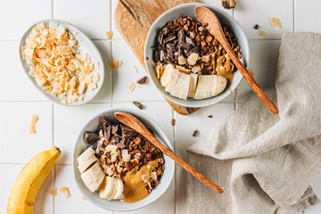 A flat lay on a white tiled table showing two bowls of chocolate granola, accompanied by chocolate pieces, banana slices, coconut chips, and yogurt for a delicious, balanced meal