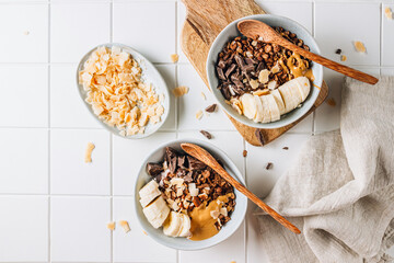 A flat lay on a white tiled table showing two bowls of chocolate granola, accompanied by chocolate pieces, banana slices, coconut chips, and yogurt for a delicious, balanced meal