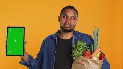 Vegan adult holding a tablet with greenscreen layout in studio, carrying paper bag full of ethically sourced chemicals free produce. Guy advocating for healthy eating with mockup screen. Camera A.