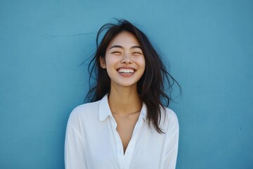 Beautiful Asian girl with long dark hair stands smiling warmly in front of a turquoise background. She is wearing a simple white shirt, exuding natural beauty and positivity.