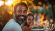 © Prostock-studio - A joyful man gazes at the camera, sitting at a beautifully set table outdoors during sunset, surrounded by friends and warm lights.