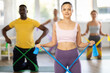 © JackF - Positive young adult Asian woman in pink top and purple leggings doing resistance band exercises with diverse group of pilates enthusiasts in light airy fitness studio