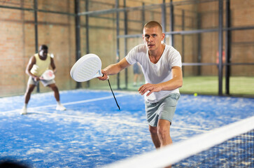 Wall Mural - Portrait of concentrated paddle tennis player preparing to hit forehand to return ball on indoor court..