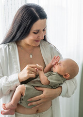 Mom stands by window and holds newborn baby in arms. Mom gently touches hand of sleeping son. Care and support, Sleep and protection. Love and relationship with an infant