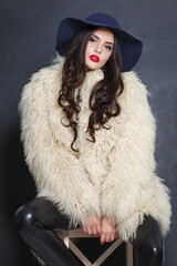 Cheerful fashion model woman in blue classic hat and white fur coat posing indoor, elegance fashion beauty portrait