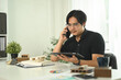 © Prathankarnpap - Young asian man talking on mobile phone at desk surrounding with several architectural models and materials