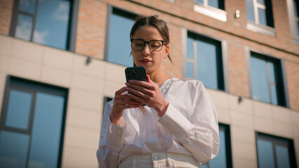 Focused Hispanic woman wearing glasses businesswoman business lady entrepreneur girl student outdoors near office building sending sms message using mobile phone search by geolocation browsing email