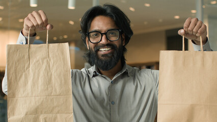 Happy joyful indian man with glasses indoors male buyer client guy customer shopper looking camera smiling showing paper bags satisfied purchases discount sale in store shows purchase goods packages